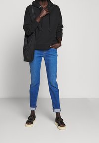 Zwarte hoodie met koordjes, blauwe broek metopgerolde pijpen en zwarte sandalen met beige zolen. Eenvoudige en casual outfit.