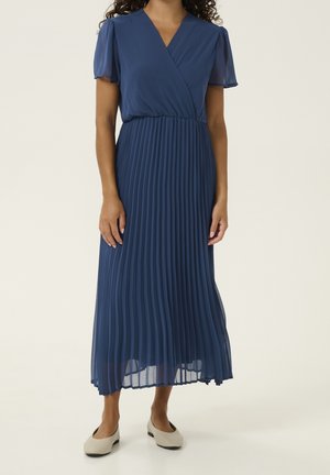 Kobieta w niebieskiej sukience z krótkim rękawem i dekoltem w serek, z plisowaną midi spódnicą, w beżowych płaskich butach, stojąca na jasnym tle.