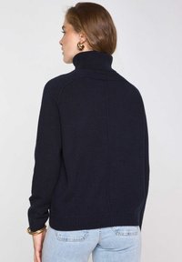 Donna con i capelli castano chiaro che indossa orecchini a cerchio dorati, un maglione a collo alto nero, jeans azzurri e bracciali dorati, di spalle rispetto alla macchina fotografica.