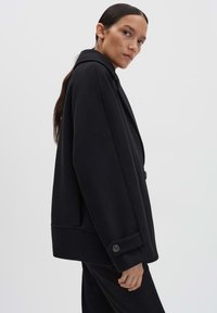 Cappotto in misto lana nero con silhouette strutturata, ampi revers e chiusura con un solo bottone. Presenta una superficie texturizzata e un dettaglio con bottone a contrasto.