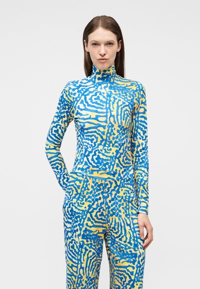 Γυναίκα με ίσια καστανά μαλλιά φοράει στενό ολόσωμο κορμάκι με μπλε και κίτρινο αφηρημένο σχέδιο, στέκεται μπροστά σε απλό φόντο.