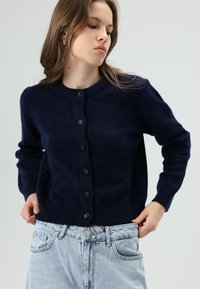 Marineblauwe gebreide cardigan met ronde halslijn, knoopsluiting aan de voorkant en geribbelde manchetten. Draagt over lichtblauwe, hooggetailleerde denim.