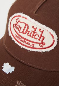 Gorra marrón de algodón, con un parche crema desgastado que lleva en rojo el texto "Von Dutch Originals". Incluye una etiqueta holográfica en el ala.