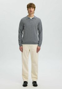 Hallikas pikkade varrukatega kootud polo särk, millel on lame krae, koos elevandiluuribade sirgete pükste ja mustade kingadega, seistes lihtsa tausta ees.