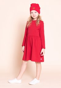 Młoda dziewczyna z kręconymi blond włosami, ubrana w czerwoną sukienkę i pasującą czerwoną czapkę, stoi z jedną nogą lekko wysuniętą do przodu i w białych trampkach.