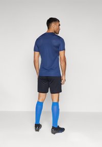 Maglietta sportiva a maniche corte blu navy, pantaloni corti neri e calze blu alte fino al ginocchio, abbinate a scarpette nere. Tessuto liscio, design aderente.