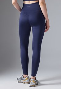 Marine aktive leggings med høy midje, glatt tekstur og subtile sømdetaljer, kombinert med sølv- og gullfargede joggesko.