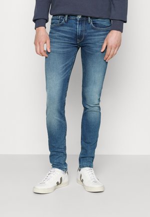 Homme portant un jean bleu slim et des baskets blanches, debout devant un fond clair uni, les mains détendues le long du corps.