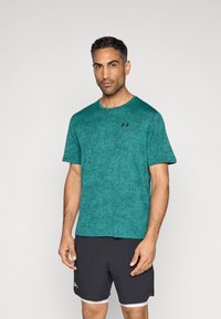 Αθλητική μπλούζα σε τιρκουάζ χρώμα με κοντά μανίκια και υφή, λαιμόκοψη στρόγγυλη, και μαύρο λογότυπο στο στήθος, σε συνδυασμό με μαύρο σορτς με λευκές λεπτομέρειες.