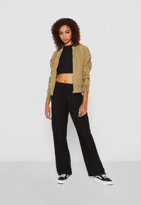 Khakifarbene Bomberjacke mit gerippten Bündchen, kombiniert mit einem schwarzen Cropped-Top und locker sitzenden schwarzen Cargo-Hosen. Schwarze Sneakers mit weißen Sohlen.