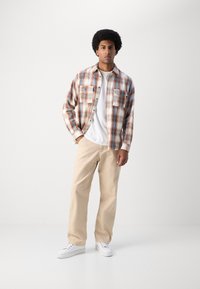 Plaid shirt in cream, brown, and blue with two chest pockets, worn over a white t-shirt and paired with beige pants and white sneakers.