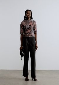Floral-patterned blouse in shades of pink and brown, paired with black high-waisted trousers. Model holds a small black bag and wears heels.