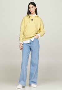 Felpa gialla chiara con coulisse e tasca frontale, abbinata a jeans larghi azzurri e sneakers bianche. Camicia bianca visibile sotto.