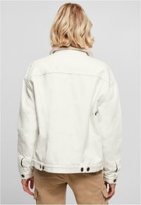 Veste en denim blanc avec avant boutonné, col doux et deux poches latérales. Coupe décontractée avec des coutures visibles et des boutons noirs.