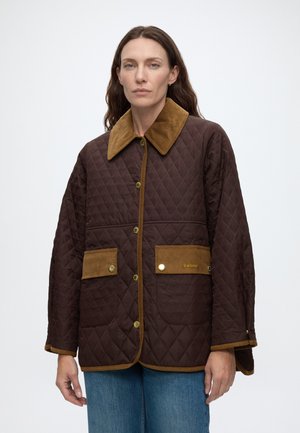 Frau trägt eine braune gesteppte Jacke mit Cordkragen und Taschen, steht vor einem schlichten hellen Hintergrund.