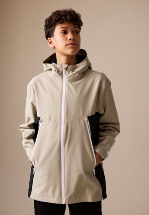 Junge Person mit kurzen lockigen Haaren trägt eine beige und schwarze Kapuzenjacke mit weißem Reißverschluss und Händen in den Taschen, schaut leicht zur Seite.