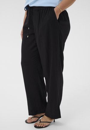 Osoba ubrana w luźne czarne spodnie ze sznurkiem, jasnoniebieską bluzkę, złote bransoletki i czarne sandały na obcasie, stojąca z jedną ręką w kieszeni.