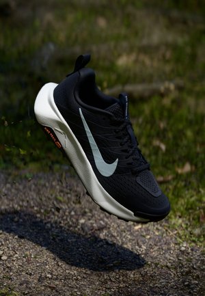 Czarno-biały but do biegania Nike unoszący się nad ziemną ścieżką z trawą w tle, pokazujący boczny profil i szczegóły podeszwy.
