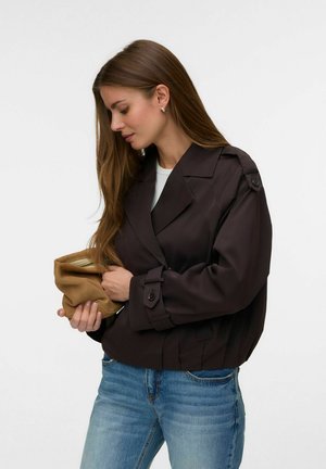 Femme aux longs cheveux bruns portant une veste marron foncé et un jean bleu, tenant et regardant une petite pochette beige sur fond blanc.