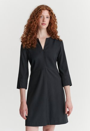 Femme aux cheveux roux bouclés portant une robe noire simple jusqu'aux genoux avec des manches trois-quarts et un décolleté en V, debout devant un fond uni.