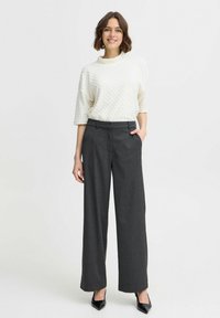 Weißer, strukturierten Pullover mit hohem Kragen, kombiniert mit dunkelgrauen weiten Hosen. Das Modell trägt schwarze spitze Absatzschuhe.