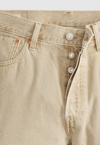 Pantalon en jean beige avec braguette à boutons partiellement ouverte, passants de ceinture visibles et rivets métalliques sur la poche avant.