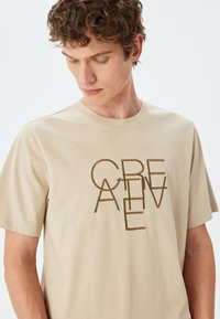 Beige bomulds t-shirt med brun, stiliseret tekstdesign. Korte ærmer og rund halsudskæring. Glat tekstur med minimal syning.