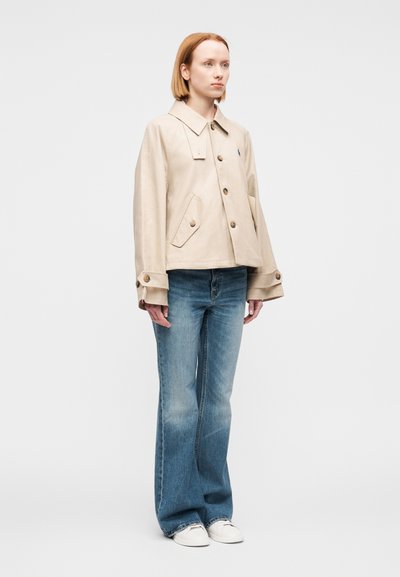 Μπεζ cropped μπουφάν με μεγάλα κουμπιά και μυτερό γιακά, συνδυασμένο με ξεθωριασμένο μπλε τζιν καμπάνα και λευκά αθλητικά παπούτσια.