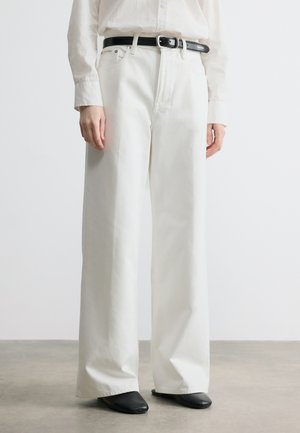 Osoba ubrana w białe spodnie z wysokim stanem i szerokimi nogawkami, czarny pasek, biała koszula zapinana na guziki oraz czarne buty z kwadratowym noskiem, stojąca na szarej podłodze.