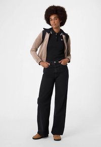 Sorte jeans med vide ben, sort poloshirt, beige og sort lynlåstrøje samt brune sko. Modellen står med hænderne på hoften og kigger fremad.