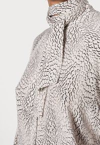 Blusa chiara con un motivo astratto nero, caratterizzata da un colletto rovesciato e un dettaglio con fiocco al collo. Il tessuto ha una consistenza liscia.