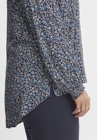 Blúzka s kvetinovým vzorom v námorníckej modrej farbe, zdobená modrými a zelenými kvetmi. Dlhé rukávy so zhromaždenými manžetami a zakriveným lemom. Hladká textúra látky.