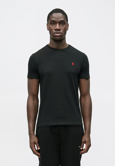 T-shirt preta de algodão com gola redonda e mangas curtas. Apresenta um pequeno logo vermelho bordado no peito esquerdo. Textura suave.