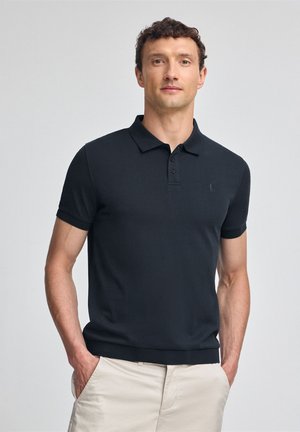 Camisa polo negra de diseño de manga corta, con un tapeta de tres botones y un sutil bordado del logo en el pecho. Combinada con pantalones cortos de color beige claro.