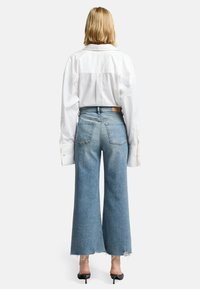 Jeans in denim chiaro a gamba larga con orlo sfrangiato, abbinati a una camicia bianca oversize. I dettagli notevoli includono tasche posteriori visibili e cuciture.
