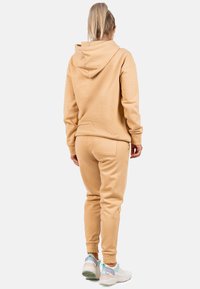 Set di felpa beige e pantaloni jogger in tessuto morbido. La felpa presenta un cappuccio con cordino, mentre i pantaloni jogger hanno polsini elastici e tasche laterali.