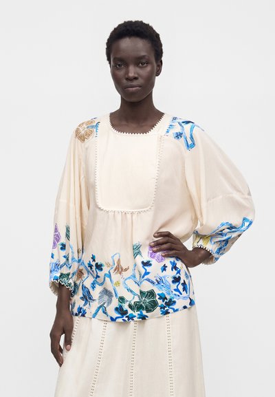 Femme aux cheveux courts portant un chemisier ample crème avec une broderie florale colorée sur les manches et l'ourlet, assorti à une jupe.