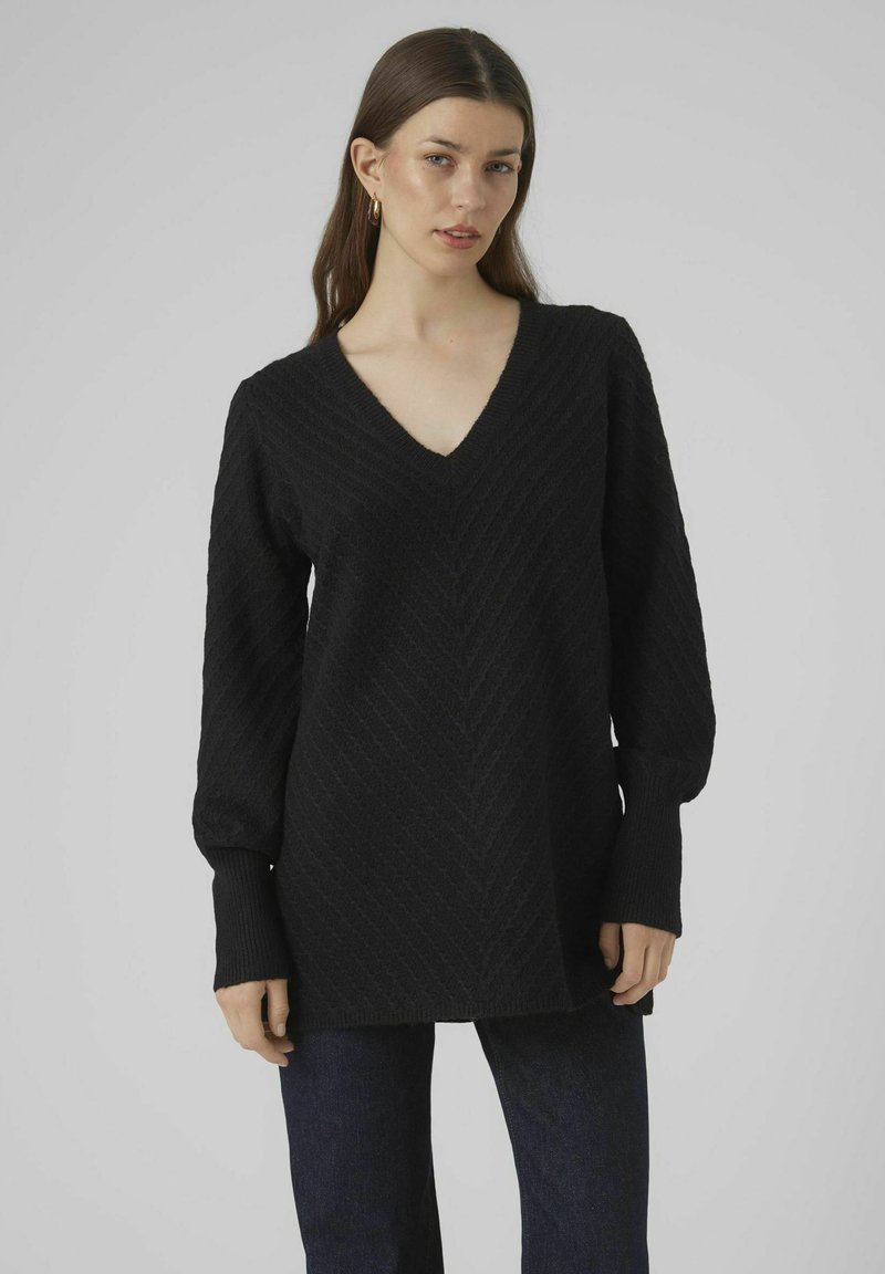 Maglione nero lavorato a maglia con scollo a V, motivo a zigzag testurizzato e maniche lunghe. Abbinato a jeans di denim blu scuro.