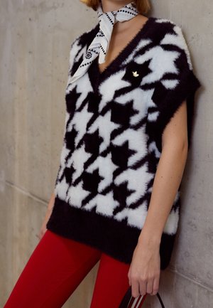 Czarna i biała futrzana kamizelka w jodełkę noszona z białą wzorzystą chustą na szyję i czerwonymi spodniami, osoba trzymająca brązową torbę.