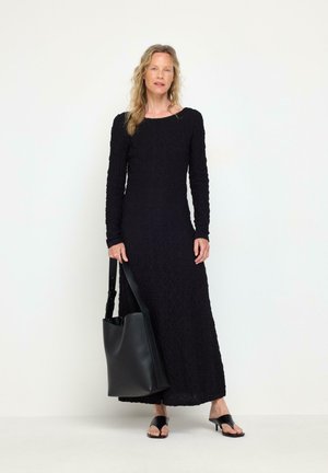 Donna con capelli lunghi e biondi che indossa un lungo vestito nero con texture e sandali neri con tacco, che tiene una grande borsa tote in pelle nera.