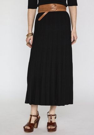 Femme portant une jupe midi plissée noire avec une large ceinture marron, des sandales à talons hauts marron, ainsi que des bracelets et bagues en or, debout devant un fond blanc.