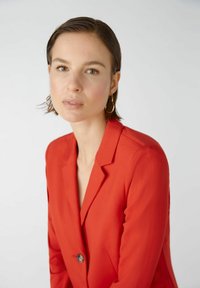 Rot maßgeschneiderter Blazer mit einem einzigen Knopf, mit Reverskragen und glatter Textur, getragen über einem V-Ausschnitt-Top. Sichtbare goldene Creolenohrringe.