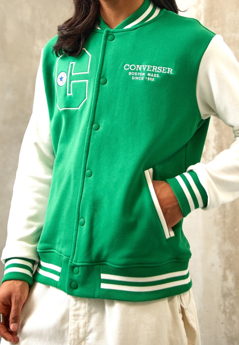 Chaqueta varsity verde y blanca con un cuerpo verde, mangas blancas, puños de canalé, un gran logo estilizado con la letra "C" y botones en la parte delantera.