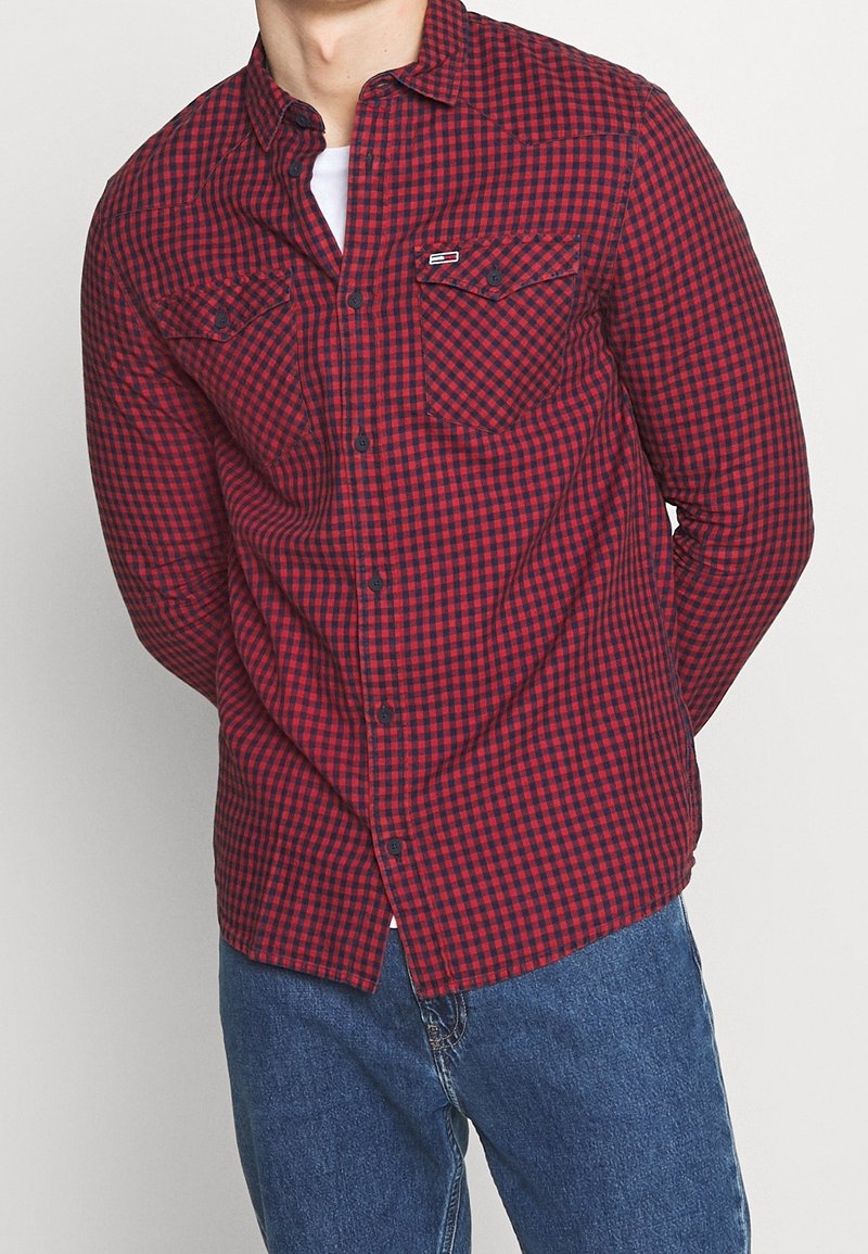 Homme portant une chemise à carreaux rouge et noir à manches longues sur un sous-maillot blanc et un jean bleu, debout avec les mains derrière le dos.