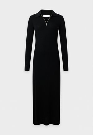 Femme portant une longue robe noire côtelée avec un col zippé, des chaussures en cuir noires et un sac à épaule noir sur un fond clair uni.