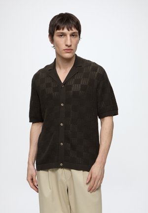 Jeune homme aux cheveux courts foncés porte une chemise en maille noire à manches courtes boutonnée et un pantalon beige sur un fond blanc uni.
