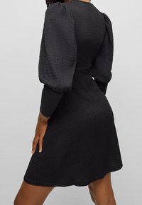 Robe noire avec manches bouffantes, tissu texturé, taille cintrée s'évasant sur les hanches, avec un col rond simple et un ourlet droit.