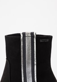 Botín de ante negro con un distintivo detalle de rayas metálicas en plata y negro en la parte posterior, con una sutil etiqueta de la marca.