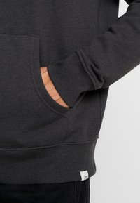 Dunkelgrauer Hoodie aus strukturiertem Stoff, mit Fronttasche, gerippten Bündchen und einem kleinen Etikett am Saum. Nahaufnahme einer Hand in der Tasche.