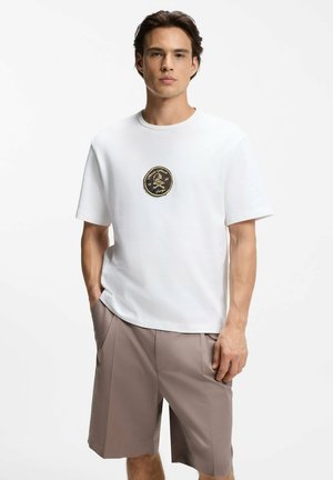 Uomo con maglietta bianca con un logo rotondo di teschio nero e oro e pantaloncini beige fino al ginocchio su sfondo bianco semplice.